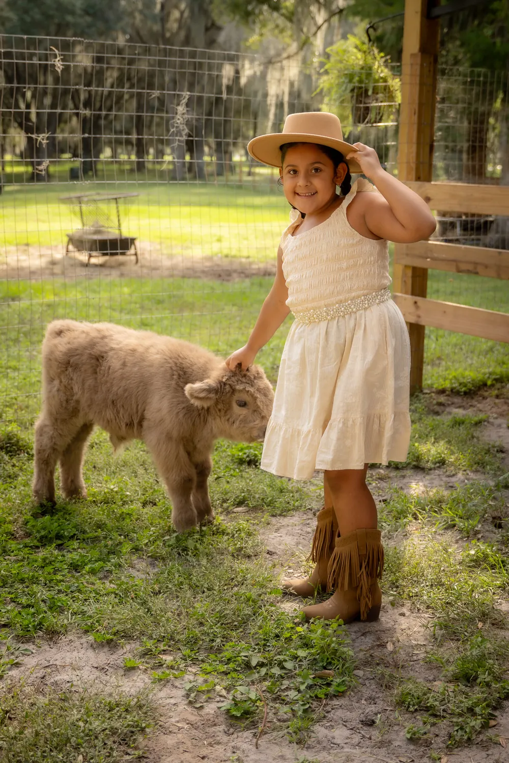 Girl in a cowboy hat petting a cream baby Highland calf